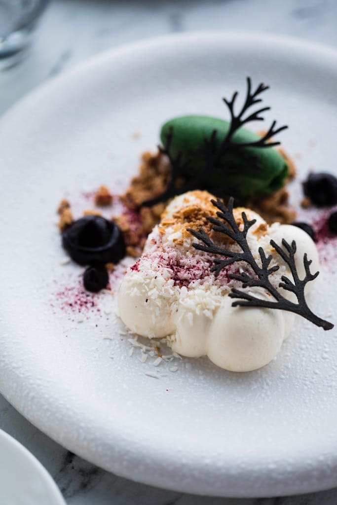 Elegant dessert with white cream, chocolate decor, and green crumble on a white plate.